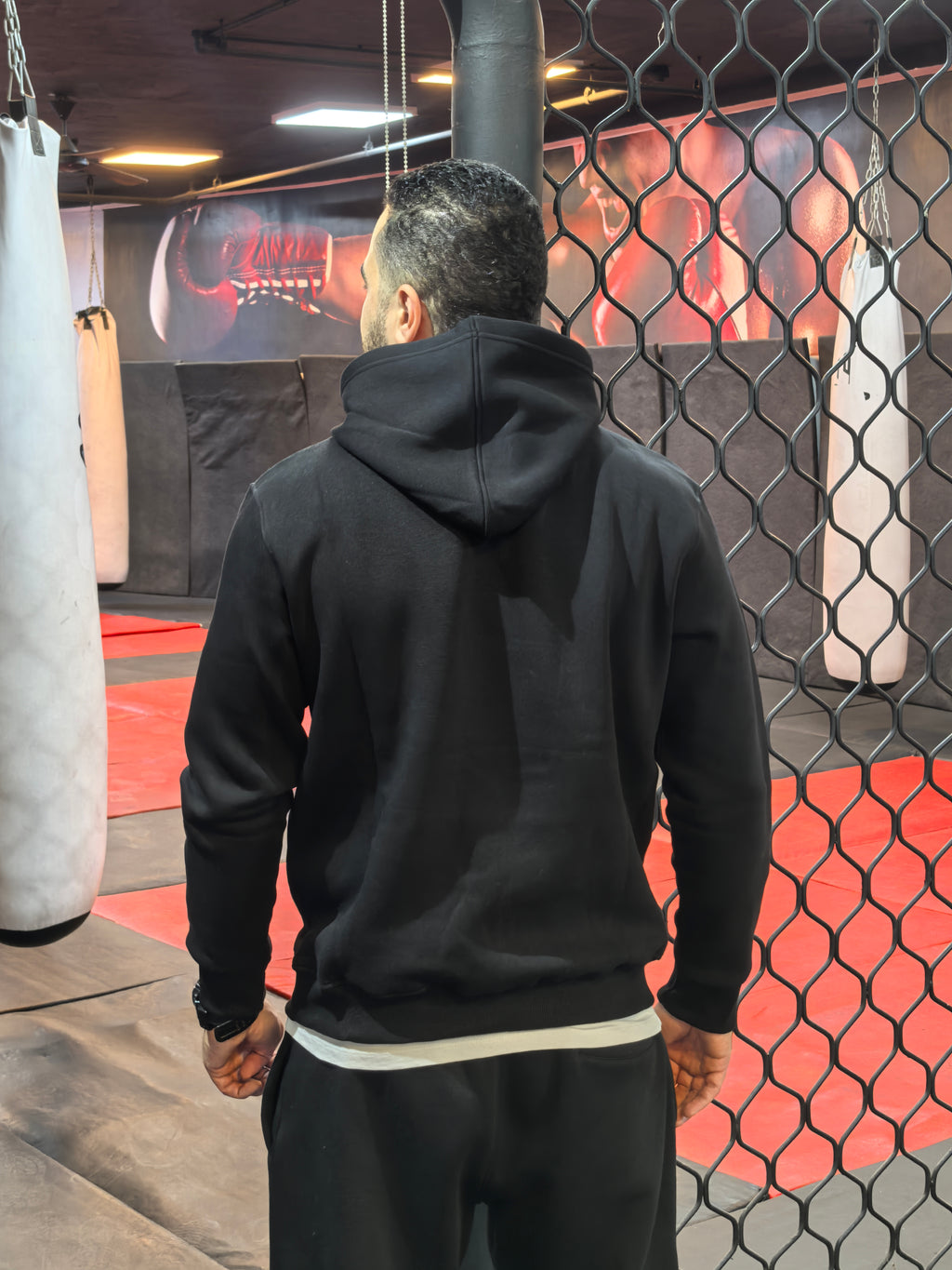 Person wearing a black hoodie in a boxing ring with punching bags.

