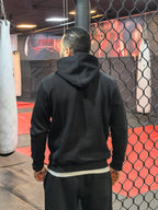 Person wearing a black hoodie in a boxing ring with punching bags.

