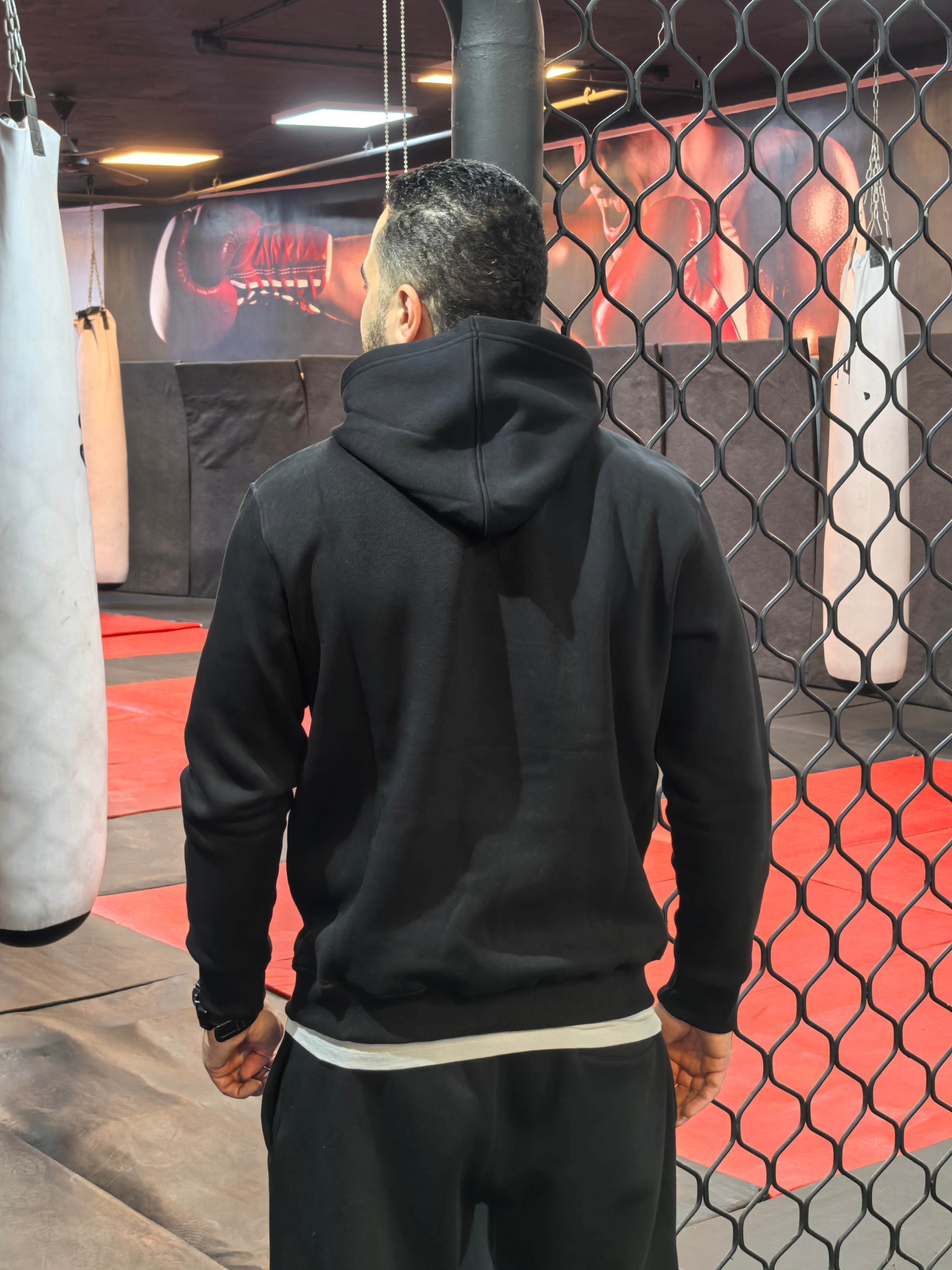 Person wearing a black hoodie in a boxing ring with punching bags.

