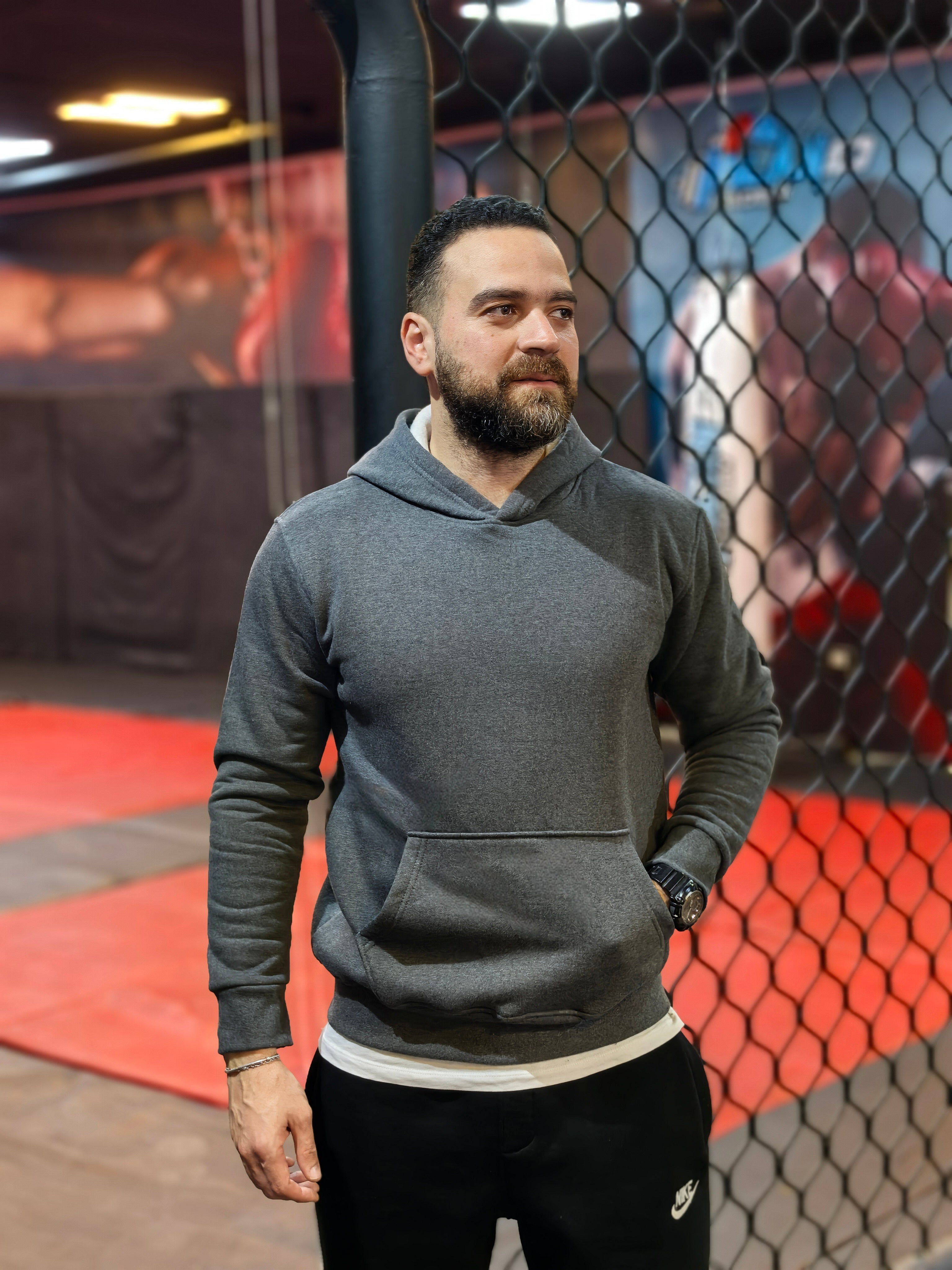 Man wearing a gray hoodie and black pants standing in front of a boxing ring.

