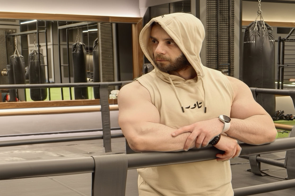 Man in a gym wearing a beige hoodie and tank top, standing by a railing.